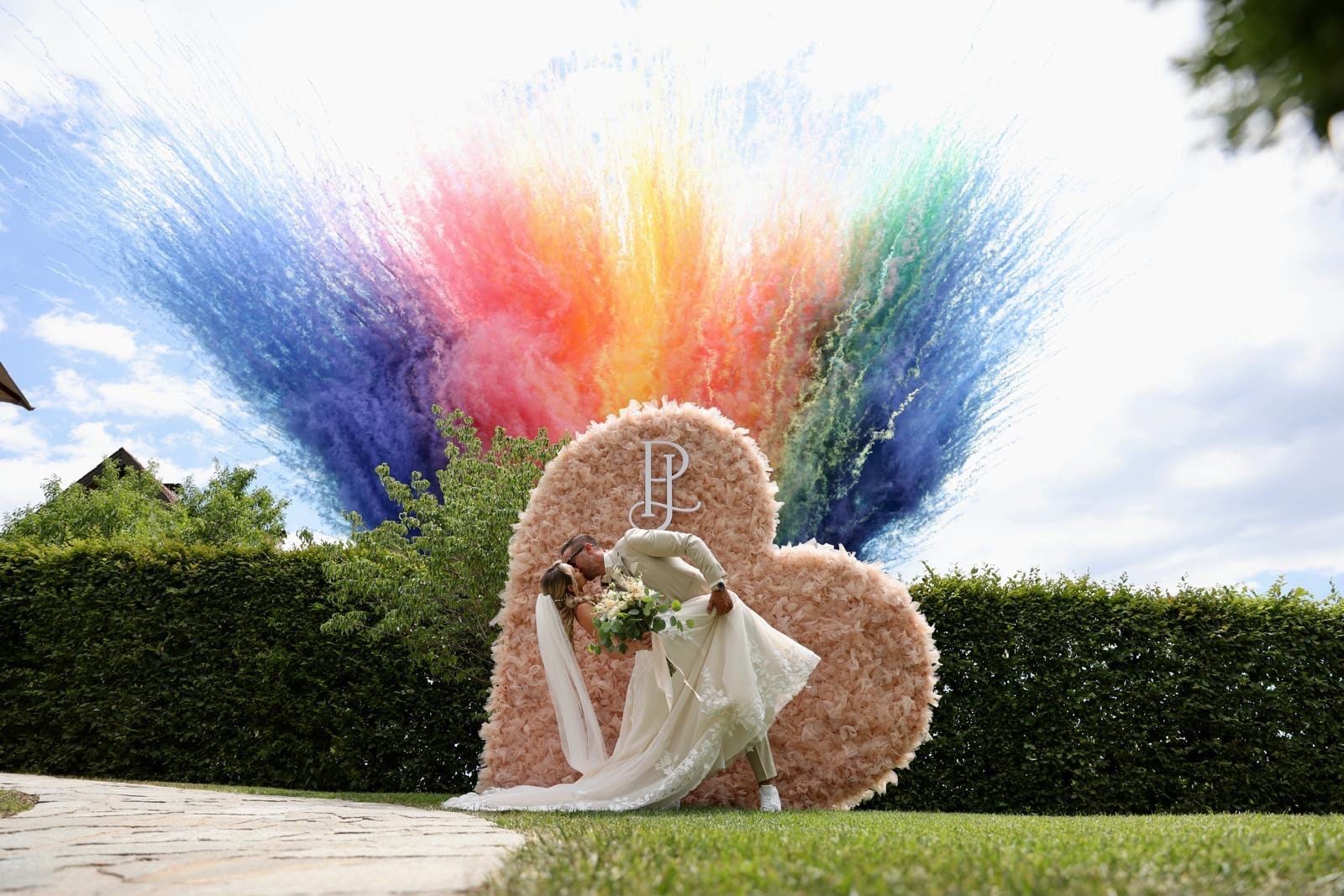 Ein Brautpaar küsst sich vor dem Herzbogen, dahinter steigt eine bunte Farbwolke auf